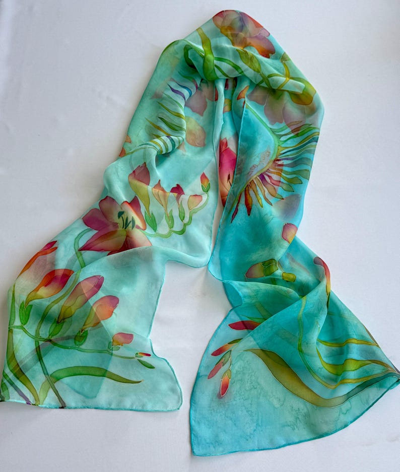Pode incluir: Um len&ccedil;o retangular de seda com um fundo turquesa vibrante e motivos florais pintados &agrave; m&atilde;o em tons de rosa, laranja e amarelo. O design inclui caules e folhas verdes delicadas sobre um tecido leve e transl&uacute;cido.