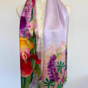 K&ouml;nnte beinhalten: Ein rechteckiger Seidenschal mit handgemaltem Blumenmuster. Eine Seite zeigt eine gro&szlig;e orange-rote Schwertlilie mit violetten Lupinen, w&auml;hrend die andere Seite in einem hellen Lavendelton gehalten ist und in violette Lupinen &uuml;bergeht.