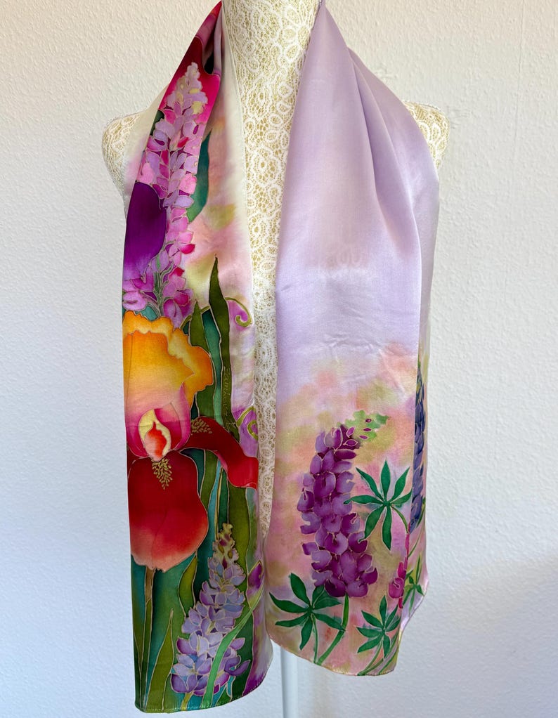 K&ouml;nnte beinhalten: Ein rechteckiger Seidenschal mit handgemaltem Blumenmuster. Eine Seite zeigt eine gro&szlig;e orange-rote Schwertlilie mit violetten Lupinen, w&auml;hrend die andere Seite in einem hellen Lavendelton gehalten ist und in violette Lupinen &uuml;bergeht.