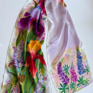 K&ouml;nnte beinhalten: Ein langer Seidenschal mit einem handgemalten Blumenmuster aus leuchtend violetten Schwertlilien, orangefarbenen Lilien sowie blauen und rosafarbenen Lupinen. Eine kleine braune Schnecke ist zwischen den gr&uuml;nen Bl&auml;ttern links zu sehen.