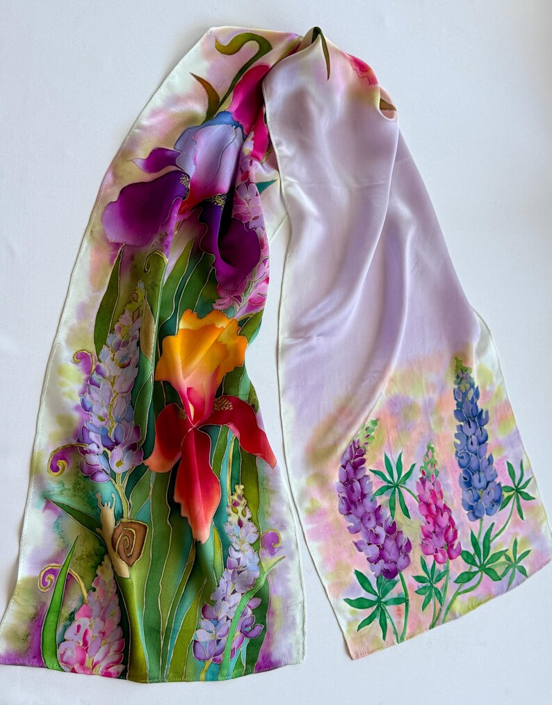 K&ouml;nnte beinhalten: Ein langer Seidenschal mit einem handgemalten Blumenmuster aus leuchtend violetten Schwertlilien, orangefarbenen Lilien sowie blauen und rosafarbenen Lupinen. Eine kleine braune Schnecke ist zwischen den gr&uuml;nen Bl&auml;ttern links zu sehen.