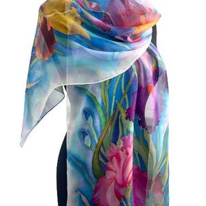May include: A lightweight silk scarf featuring a hand-painted floral design with vibrant iris flowers in shades of pink, purple, and yellow. The background consists of flowing blue and white watercolor-style washes with green stems and leaves.