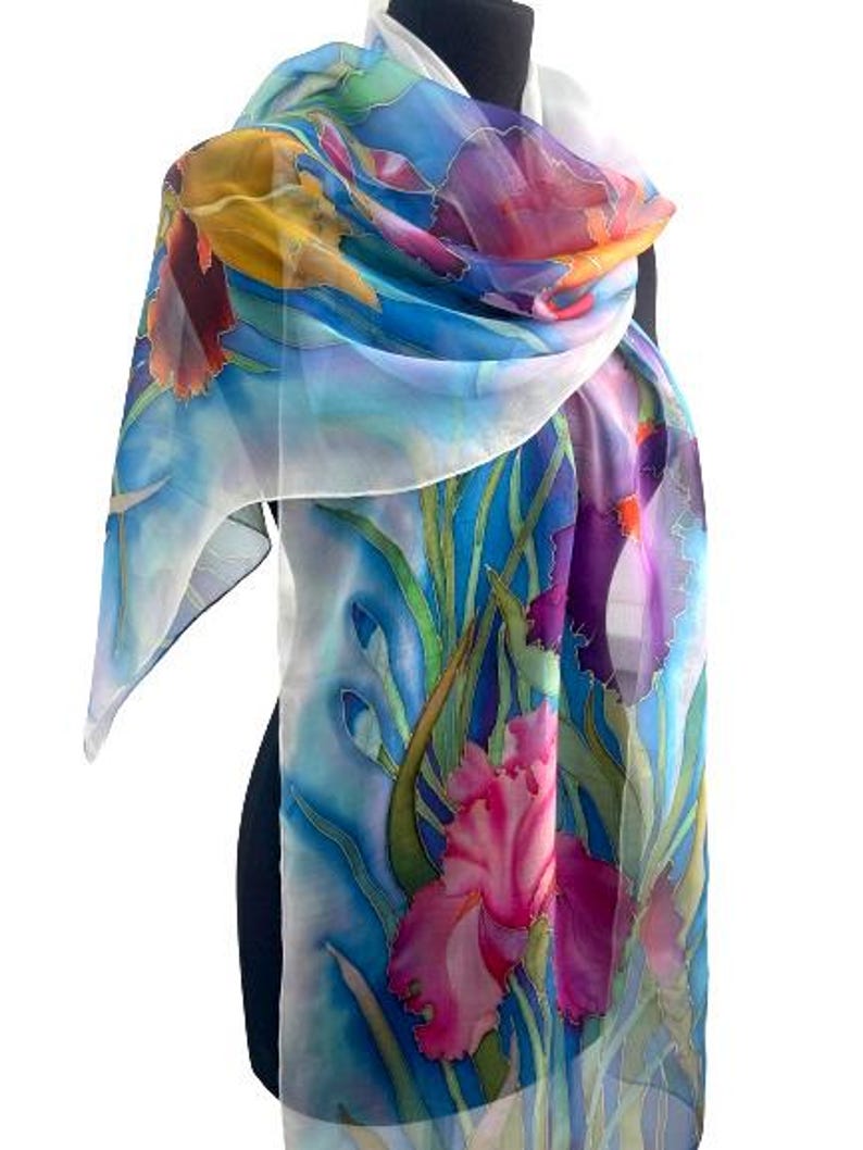 May include: A lightweight silk scarf featuring a hand-painted floral design with vibrant iris flowers in shades of pink, purple, and yellow. The background consists of flowing blue and white watercolor-style washes with green stems and leaves.