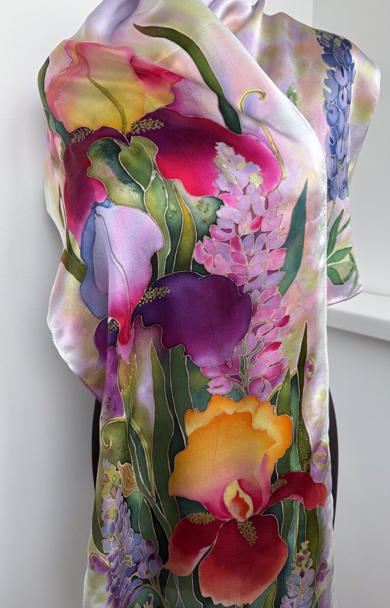 K&ouml;nnte beinhalten: Ein Seidenschal mit handgemaltem Blumenmuster, das Schwertlilien und Lupinen in Violett-, Rosa-, Orange- und Gelbt&ouml;nen zeigt. Der Stoff hat eine glatte, gl&auml;nzende Oberfl&auml;che mit gr&uuml;nen Blattakzenten und dem Namenszug Ramkowska.