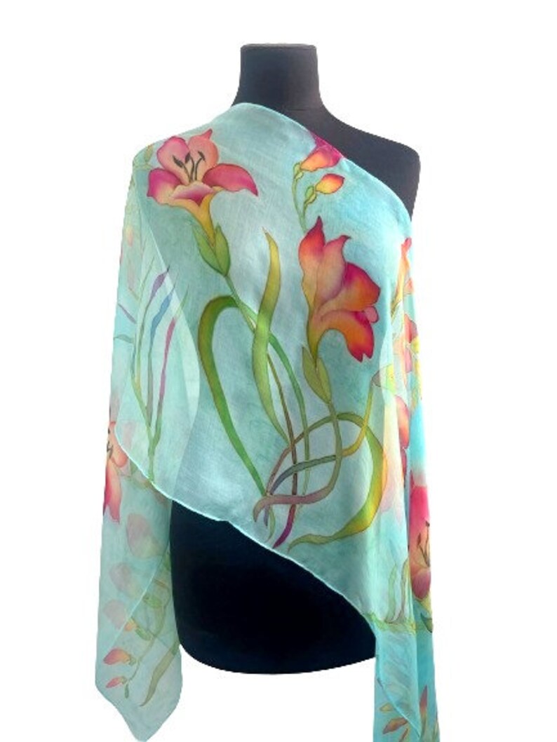 Pode incluir: Um len&ccedil;o leve e semitransparente com fundo turquesa claro e um padr&atilde;o floral pintado &agrave; m&atilde;o. O design apresenta flores vibrantes em tons de rosa e laranja, semelhantes a l&iacute;rios, com caules e folhas verdes longos, drapeado num manequim escuro.