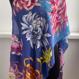 May include: A blue silk scarf with a floral pattern of pink, white, and yellow chrysanthemums. The scarf is hand-painted with the artist's signature in the upper right corner.