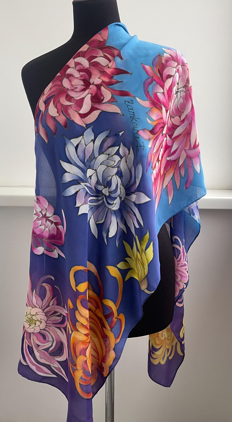 May include: A blue silk scarf with a floral pattern of pink, white, and yellow chrysanthemums. The scarf is hand-painted with the artist's signature in the upper right corner.