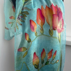 Pode incluir: Um len&ccedil;o leve e semitransparente num tom azul turquesa vibrante, com um design floral pintado &agrave; m&atilde;o. O padr&atilde;o apresenta flores alongadas em forma de trombeta com gradientes de rosa, laranja e amarelo, acompanhadas por caules verdes.