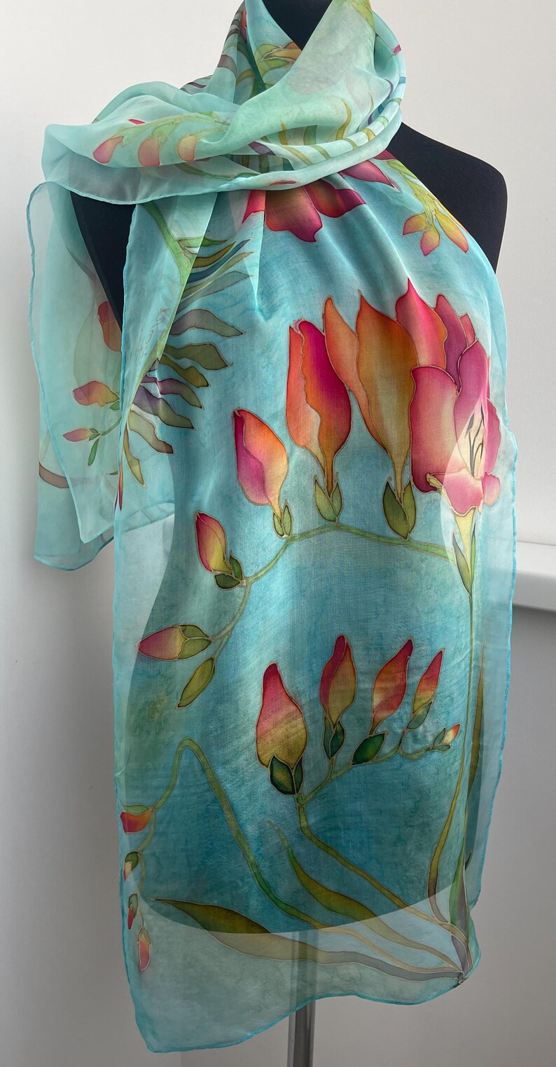 Pode incluir: Um len&ccedil;o leve e semitransparente num tom azul turquesa vibrante, com um design floral pintado &agrave; m&atilde;o. O padr&atilde;o apresenta flores alongadas em forma de trombeta com gradientes de rosa, laranja e amarelo, acompanhadas por caules verdes.