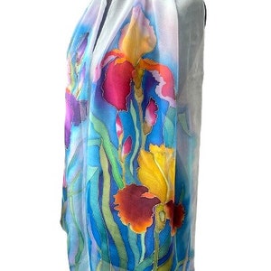 May include: A lightweight scarf featuring a hand-painted floral design of vibrant irises in shades of deep red, bright yellow, and purple. The flowers are set against a background of flowing green stems and leaves, with a soft white border.