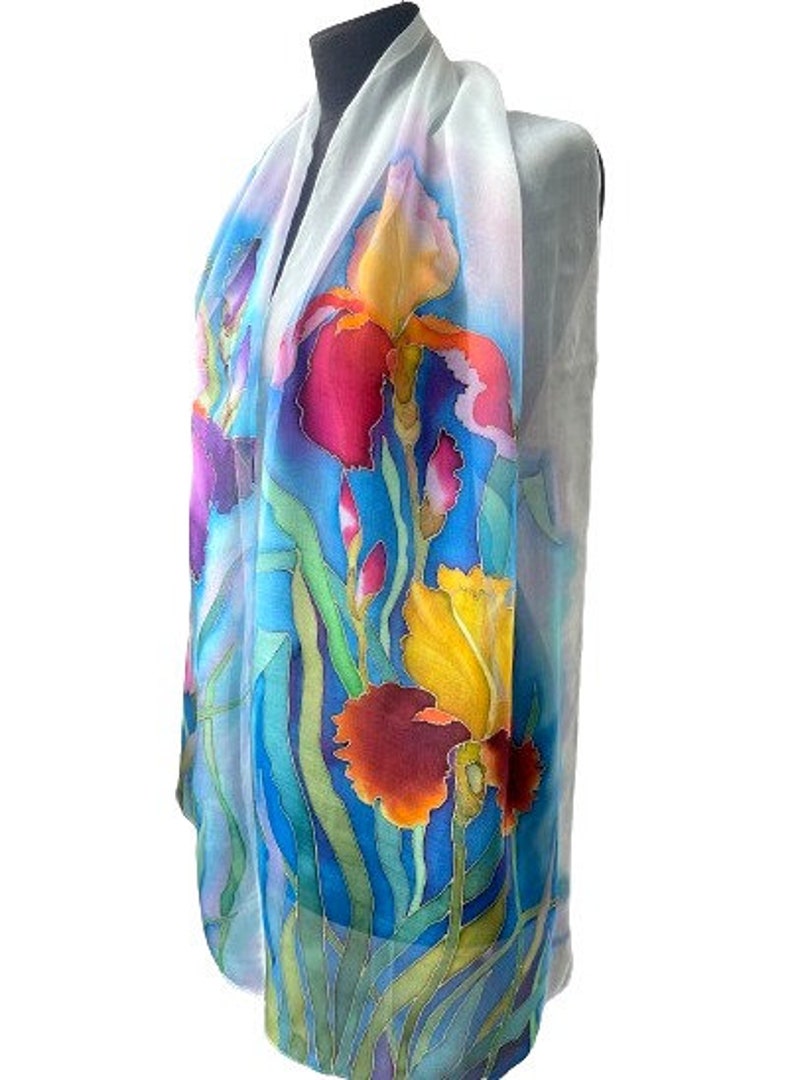 May include: A lightweight scarf featuring a hand-painted floral design of vibrant irises in shades of deep red, bright yellow, and purple. The flowers are set against a background of flowing green stems and leaves, with a soft white border.
