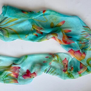 Pode incluir: Um len&ccedil;o retangular leve e semitransparente num tom azul turquesa vibrante. O tecido apresenta um design bot&acirc;nico pintado &agrave; m&atilde;o com folhas verdes fluidas e flores delicadas e multicoloridas em tons de rosa, laranja e amarelo.