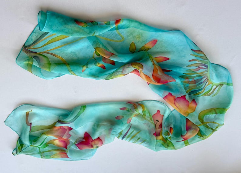 Pode incluir: Um len&ccedil;o retangular leve e semitransparente num tom azul turquesa vibrante. O tecido apresenta um design bot&acirc;nico pintado &agrave; m&atilde;o com folhas verdes fluidas e flores delicadas e multicoloridas em tons de rosa, laranja e amarelo.