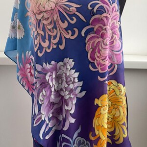 May include: A blue silk scarf with a gradient pattern of flowers. The flowers are pink, purple, yellow, and blue. The scarf is draped over a mannequin.
