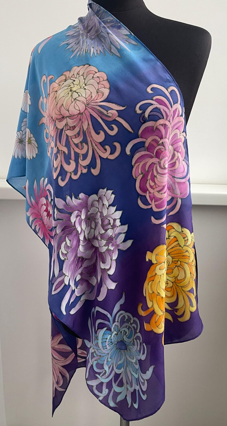 May include: A blue silk scarf with a gradient pattern of flowers. The flowers are pink, purple, yellow, and blue. The scarf is draped over a mannequin.