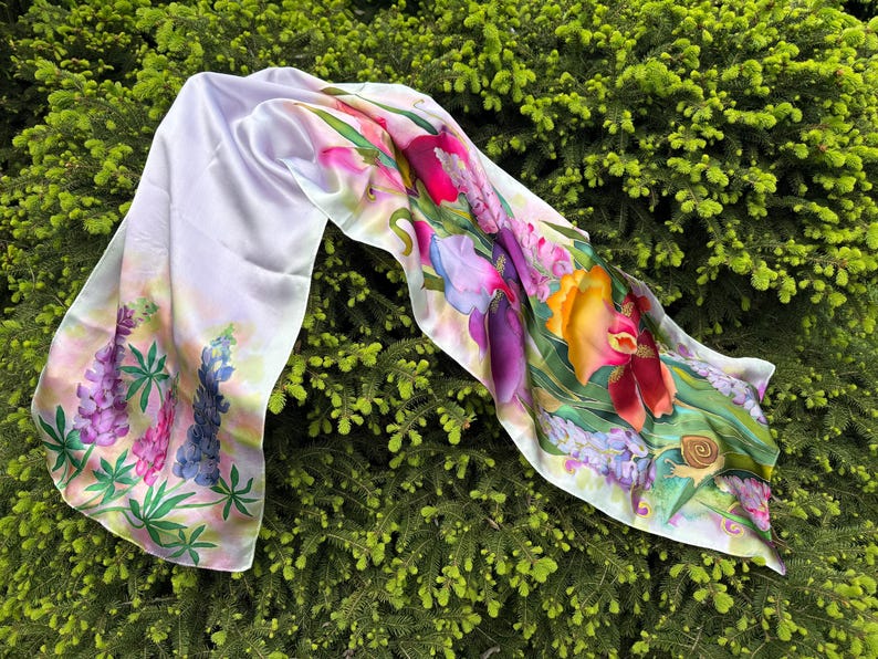K&ouml;nnte beinhalten: Ein rechteckiger Seidenschal mit einem lebendigen botanischen Druck, der violette und rosafarbene Lupinen, gro&szlig;e gelbe und rote Irisbl&uuml;ten sowie eine kleine braune Schnecke auf wei&szlig;em Grund mit gr&uuml;nen Blattmotiven zeigt.