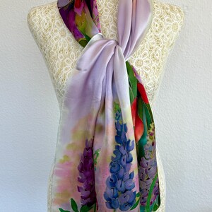 K&ouml;nnte beinhalten: Ein rechteckiger Seidenschal mit einem handgemalten Blumenmuster aus Lupinen in Violett-, Blau- und Rosat&ouml;nen. Der Schal hat eine weiche, glatte Textur und ist so drapiert, dass ein farbenfrohes botanisches Muster mit gr&uuml;nen Bl&auml;ttern sichtbar wird.