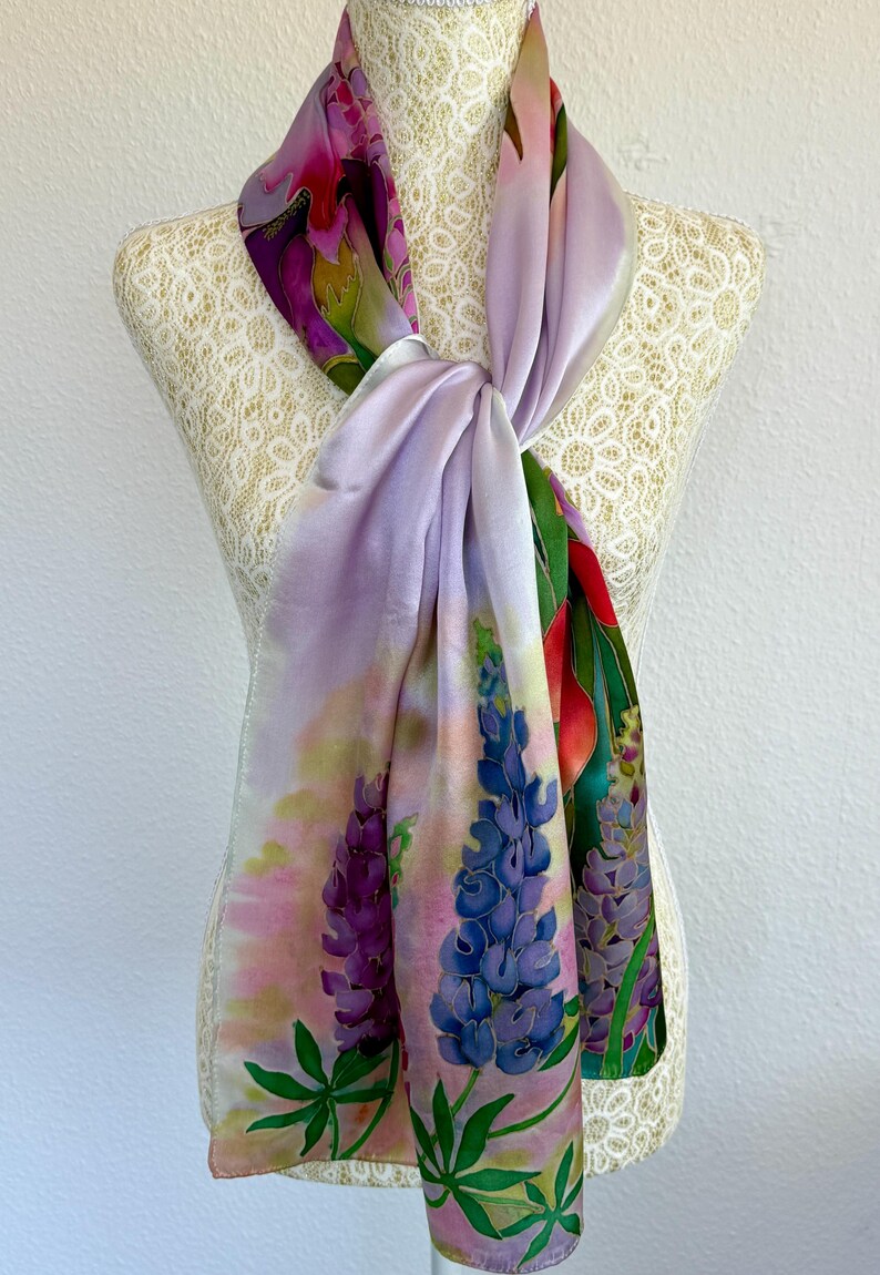 K&ouml;nnte beinhalten: Ein rechteckiger Seidenschal mit einem handgemalten Blumenmuster aus Lupinen in Violett-, Blau- und Rosat&ouml;nen. Der Schal hat eine weiche, glatte Textur und ist so drapiert, dass ein farbenfrohes botanisches Muster mit gr&uuml;nen Bl&auml;ttern sichtbar wird.
