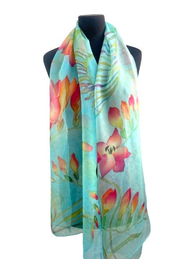 Pode incluir: Um len&ccedil;o leve com um padr&atilde;o floral vibrante com flores em tons de rosa, laranja e amarelo sobre um fundo azul turquesa suave. O design inclui caules e folhas verdes delicadas, criando um motivo bot&acirc;nico.