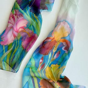 May include: A long, rectangular silk scarf featuring a hand-painted floral design of irises in shades of deep purple, vibrant pink, golden yellow, and orange. The flowers are set against a background of blue and green foliage on a white base.