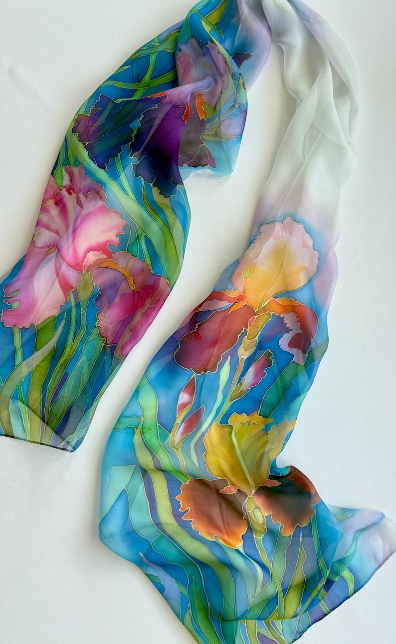 May include: A long, rectangular silk scarf featuring a hand-painted floral design of irises in shades of deep purple, vibrant pink, golden yellow, and orange. The flowers are set against a background of blue and green foliage on a white base.