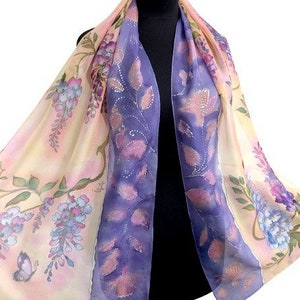 Puede incluir: Una bufanda de seda con un diseño floral en tonos morado, rosa y amarillo. La bufanda presenta un patrón de glicinas con detalles de mariposas y hojas. La bufanda está drapeada sobre un maniquí negro.