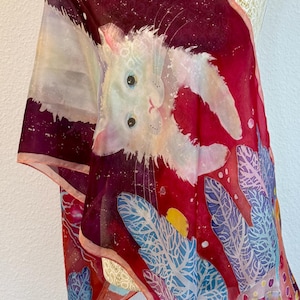 Pode incluir: Um lenço leve com fundo vermelho profundo, uma ilustração de um gato branco, detalhes de manchas brancas e padrões de folhas em azul e roxo. O tecido apresenta um design artístico vibrante com um caimento suave e fluido.