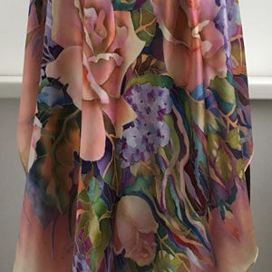 May include: A silk scarf featuring a hand-painted floral pattern with large pink roses, clusters of small purple flowers, and green foliage. The design incorporates blended shades of magenta, violet, and soft peach across the fabric surface.