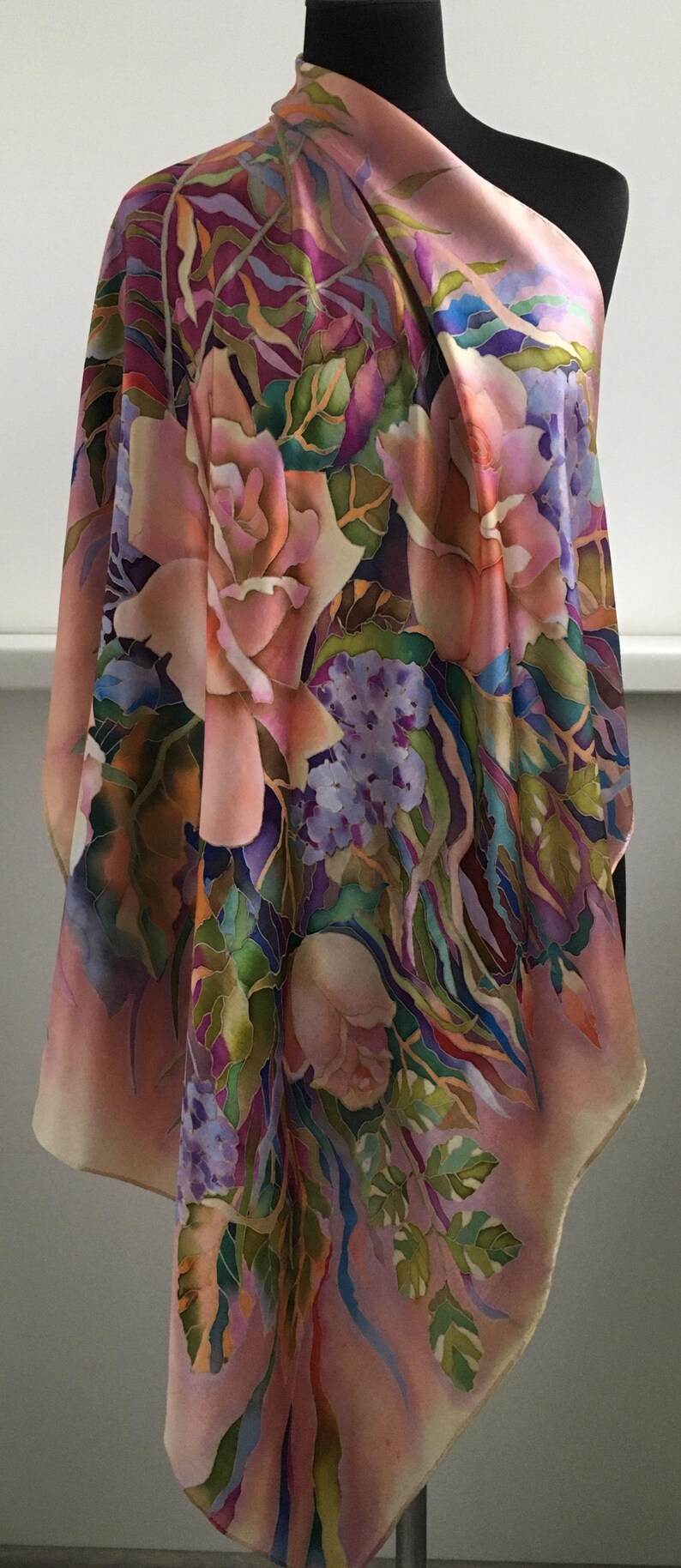 May include: A silk scarf featuring a hand-painted floral pattern with large pink roses, clusters of small purple flowers, and green foliage. The design incorporates blended shades of magenta, violet, and soft peach across the fabric surface.