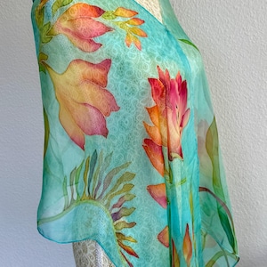Pode incluir: Um len&ccedil;o de seda leve e semitransparente com um fundo turquesa vibrante e um padr&atilde;o subtil e repetitivo semelhante a renda. O len&ccedil;o &eacute; decorado com motivos florais pintados &agrave; m&atilde;o em grande escala em tons de laranja, rosa e amarelo.