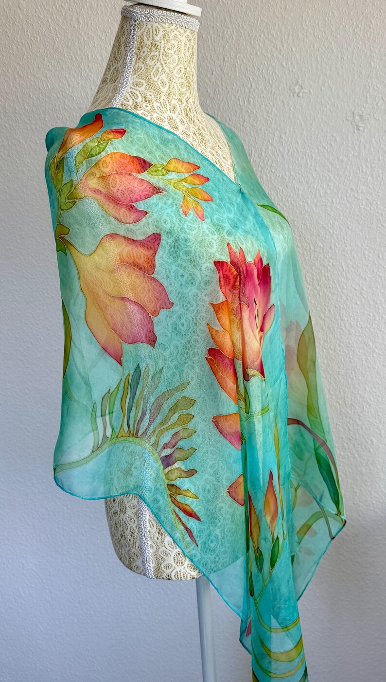 Pode incluir: Um len&ccedil;o de seda leve e semitransparente com um fundo turquesa vibrante e um padr&atilde;o subtil e repetitivo semelhante a renda. O len&ccedil;o &eacute; decorado com motivos florais pintados &agrave; m&atilde;o em grande escala em tons de laranja, rosa e amarelo.