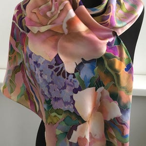 May include: A square silk scarf featuring a hand-painted floral pattern with large pink roses, clusters of purple hydrangeas, and green foliage. The design uses soft gradients of peach, lavender, and deep violet, with dark outlines defining the petals.
