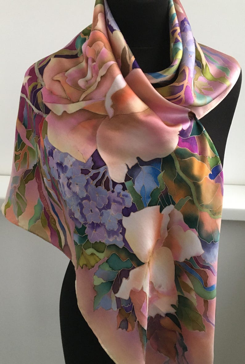 May include: A square silk scarf featuring a hand-painted floral pattern with large pink roses, clusters of purple hydrangeas, and green foliage. The design uses soft gradients of peach, lavender, and deep violet, with dark outlines defining the petals.