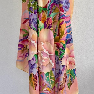 May include: A rectangular silk scarf featuring a hand-painted floral pattern with large pink roses, clusters of purple hydrangeas, and green foliage. The background is a soft peach color, and the design includes vibrant accents of blue and yellow.