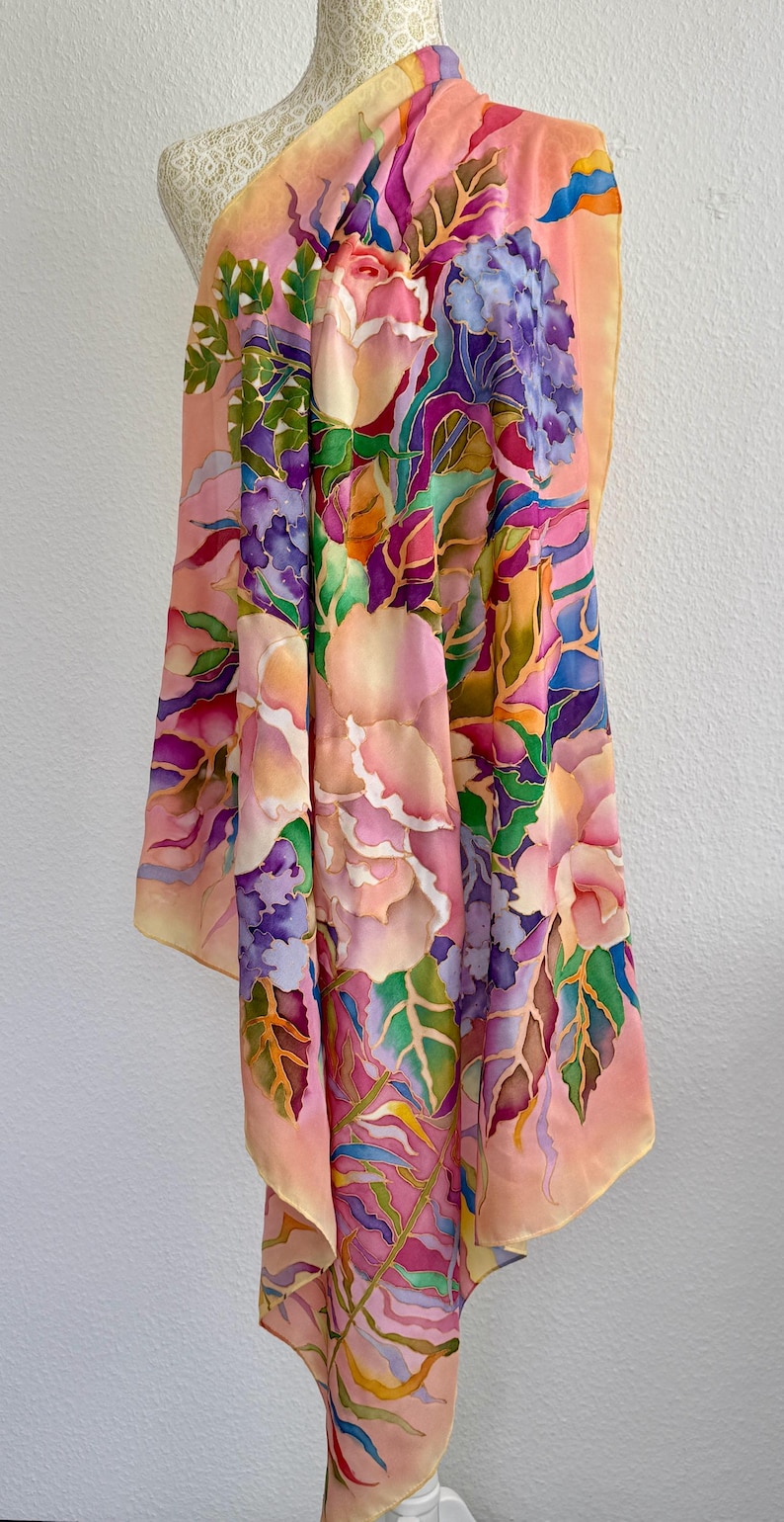 May include: A rectangular silk scarf featuring a hand-painted floral pattern with large pink roses, clusters of purple hydrangeas, and green foliage. The background is a soft peach color, and the design includes vibrant accents of blue and yellow.