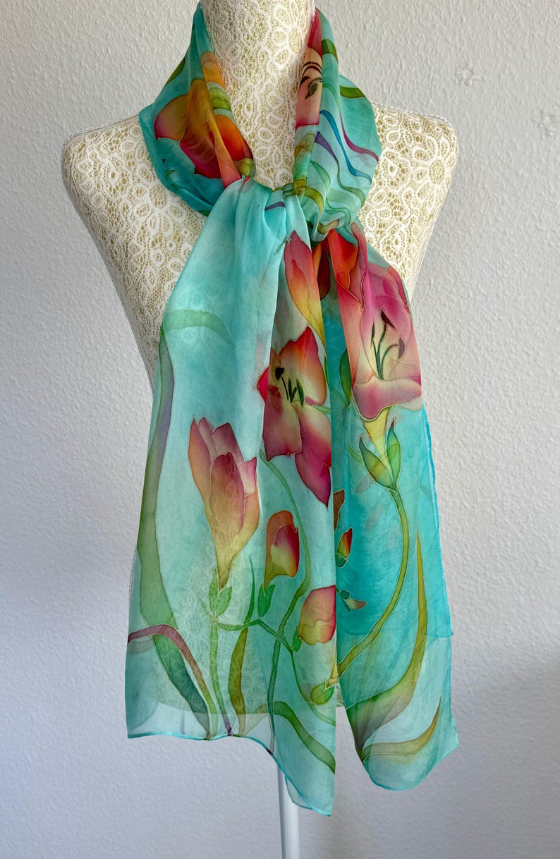 Pode incluir: Um len&ccedil;o leve com um fundo turquesa vibrante e motivos florais pintados &agrave; m&atilde;o em tons de rosa, laranja e amarelo. O design apresenta caules e folhas verdes alongadas que se entrela&ccedil;am pelo tecido, criando um padr&atilde;o bot&acirc;nico fluido.