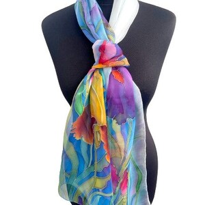 May include: A rectangular silk scarf featuring a vibrant hand-painted floral pattern in shades of blue, purple, yellow, orange, and green. The scarf is tied in a knot at the neck, displaying a white section near the top and flowing fabric below.