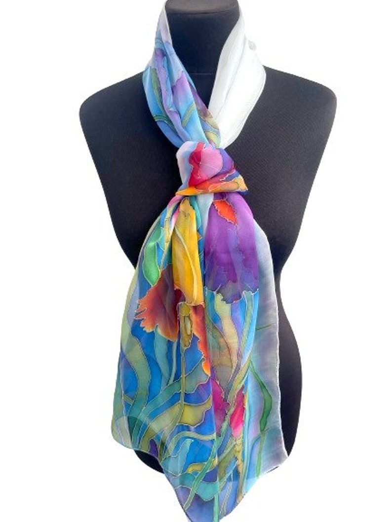 May include: A rectangular silk scarf featuring a vibrant hand-painted floral pattern in shades of blue, purple, yellow, orange, and green. The scarf is tied in a knot at the neck, displaying a white section near the top and flowing fabric below.
