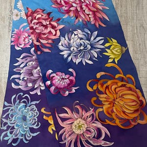 May include: A silk scarf with a blue and purple gradient background featuring a hand-painted design of colorful chrysanthemums. The scarf is signed "Zarina" in the top right corner.