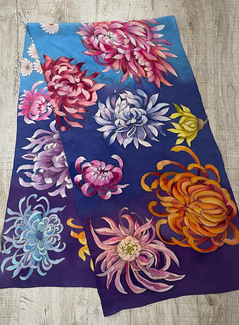 May include: A silk scarf with a blue and purple gradient background featuring a hand-painted design of colorful chrysanthemums. The scarf is signed "Zarina" in the top right corner.