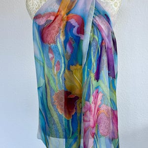 May include: A lightweight, semi-sheer silk scarf featuring a hand-painted floral design of vibrant irises in shades of deep red, magenta, yellow, and purple. The background is a gradient of bright blue and green, with long, slender green leaves.
