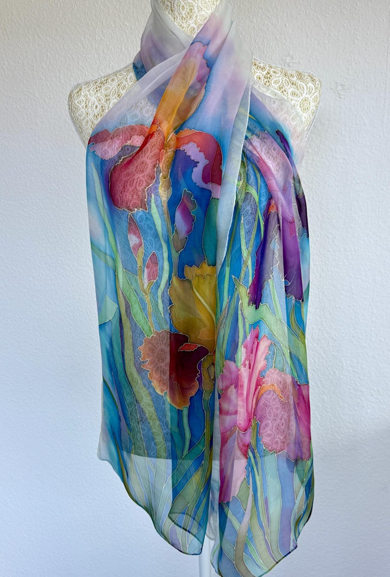 May include: A lightweight, semi-sheer silk scarf featuring a hand-painted floral design of vibrant irises in shades of deep red, magenta, yellow, and purple. The background is a gradient of bright blue and green, with long, slender green leaves.