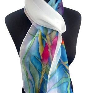 May include: A lightweight scarf featuring a hand-painted botanical design with vibrant pink, green, and yellow floral motifs set against a bright blue background. The scarf has a white border and a sheer, flowing texture draped around the neck.
