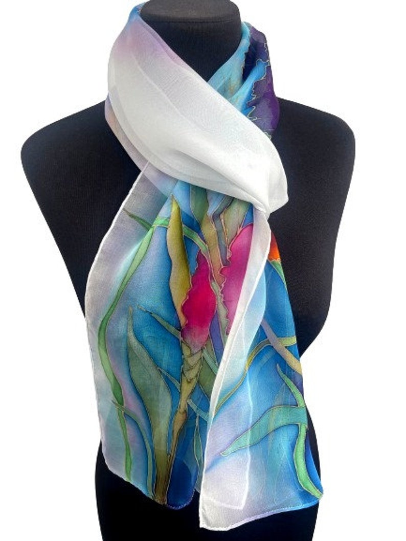 May include: A lightweight scarf featuring a hand-painted botanical design with vibrant pink, green, and yellow floral motifs set against a bright blue background. The scarf has a white border and a sheer, flowing texture draped around the neck.