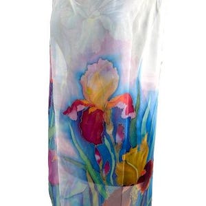 May include: A sleeveless dress featuring a hand-painted floral design on a white background. The garment displays vibrant iris flowers in shades of deep red, yellow, and purple, set against a backdrop of blue and green foliage with a soft watercolor effect.