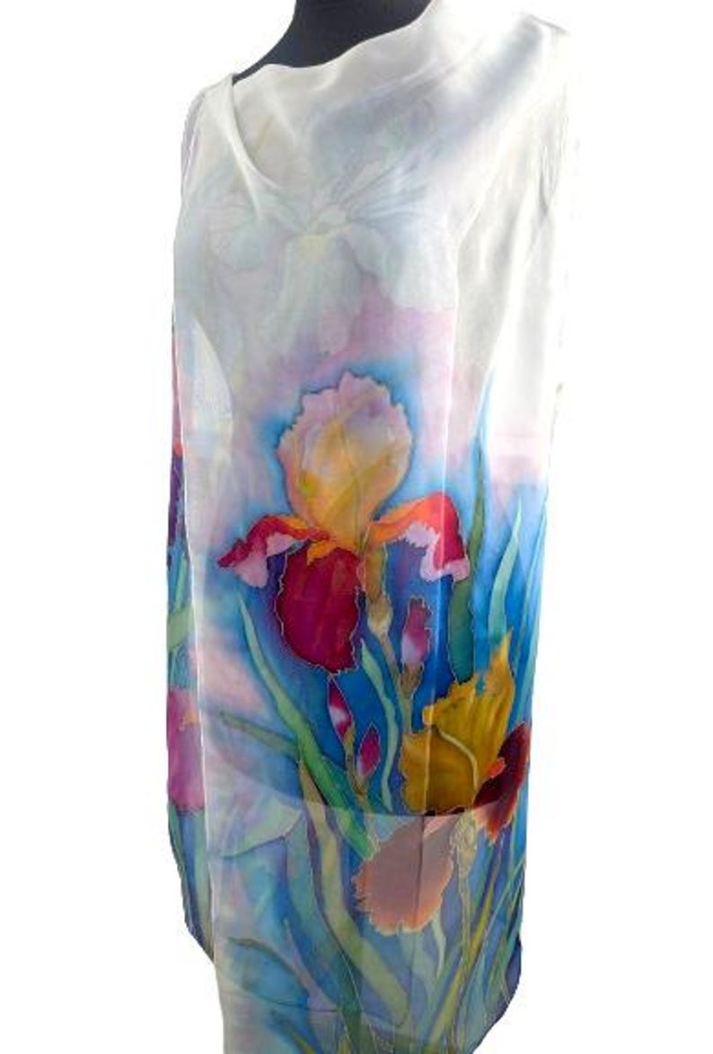 May include: A sleeveless dress featuring a hand-painted floral design on a white background. The garment displays vibrant iris flowers in shades of deep red, yellow, and purple, set against a backdrop of blue and green foliage with a soft watercolor effect.