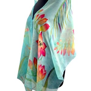 Pode incluir: Um len&ccedil;o transparente azul-claro com um design floral pintado &agrave; m&atilde;o, apresentando flores vibrantes em vermelho, laranja e amarelo, juntamente com folhagem verde. O tecido leve est&aacute; drapeado num manequim, destacando a transpar&ecirc;ncia e o padr&atilde;o bot&acirc;nico.