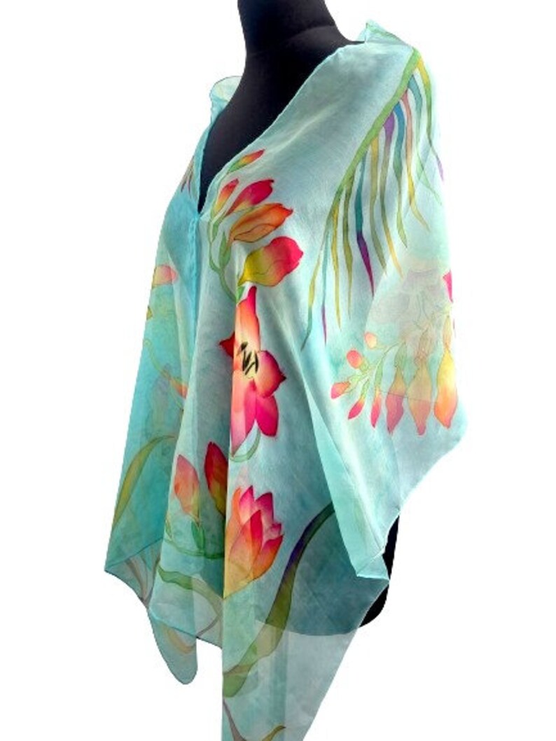 Pode incluir: Um len&ccedil;o transparente azul-claro com um design floral pintado &agrave; m&atilde;o, apresentando flores vibrantes em vermelho, laranja e amarelo, juntamente com folhagem verde. O tecido leve est&aacute; drapeado num manequim, destacando a transpar&ecirc;ncia e o padr&atilde;o bot&acirc;nico.