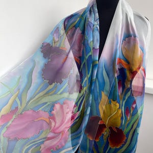 May include: A lightweight silk scarf featuring a hand-painted floral pattern of irises in shades of pink, purple, and yellow against a light blue background with green stems and leaves. The fabric is translucent and drapes over a black mannequin.