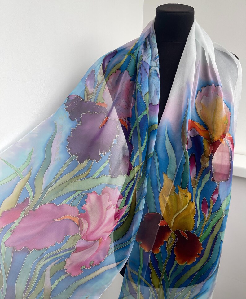 May include: A lightweight silk scarf featuring a hand-painted floral pattern of irises in shades of pink, purple, and yellow against a light blue background with green stems and leaves. The fabric is translucent and drapes over a black mannequin.